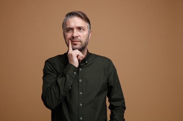Uomo di mezza età con i capelli grigi in camicia di colore scuro che guarda da parte pensando perplesso tenendo il dito sul mento in piedi su sfondo marrone