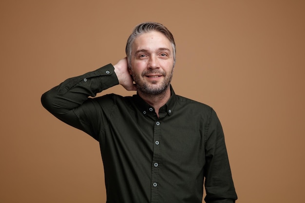 Uomo di mezza età con i capelli grigi in camicia di colore scuro che guarda l'obbiettivo felice e soddisfatto tenendo la mano dietro la testa rilassato e sorridente positivo in piedi su sfondo marrone