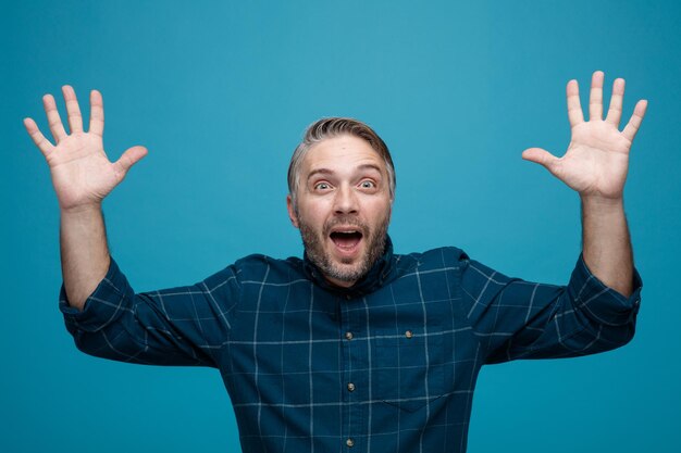 Uomo di mezza età con i capelli grigi in camicia di colore scuro che guarda la fotocamera felice ed eccitato alzando le braccia in segno di resa in piedi su sfondo blu