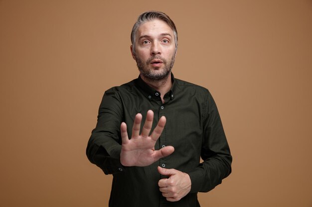 Uomo di mezza età con i capelli grigi in camicia di colore scuro che guarda la telecamera preoccupato di fare un gesto di arresto con la mano in piedi su sfondo marrone