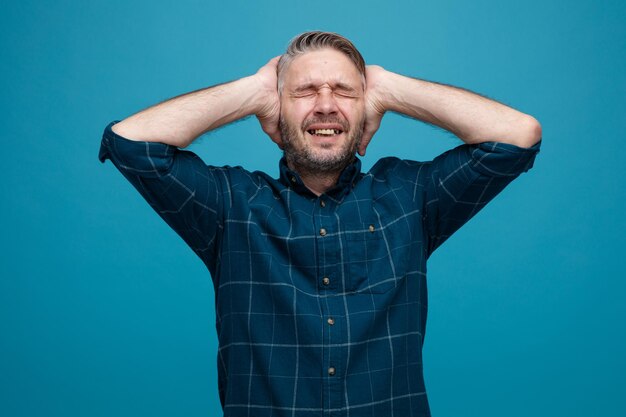 Uomo di mezza età con i capelli grigi in camicia di colore scuro che sembra infastidito e stressato tenendo le mani sulla testa in piedi su sfondo blu