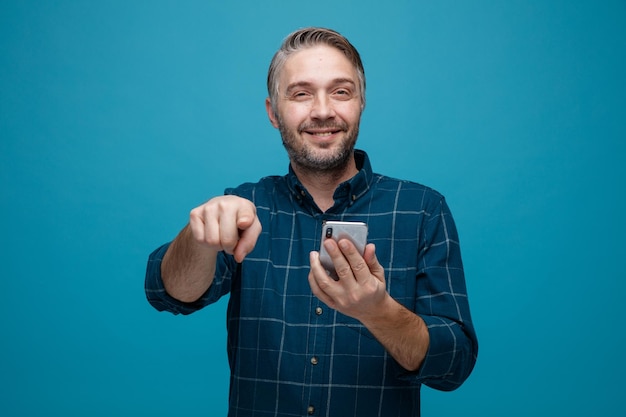 Uomo di mezza età con i capelli grigi in camicia di colore scuro che tiene lo smartphone puntato con il dito indice alla telecamera sorridente fiducioso felice e positivo in piedi su sfondo blu
