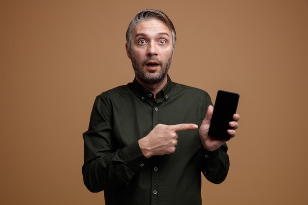 Uomo di mezza età con i capelli grigi in camicia di colore scuro che tiene lo smartphone puntato con il dito indice mentre è stupito e sorpreso in piedi su sfondo marrone