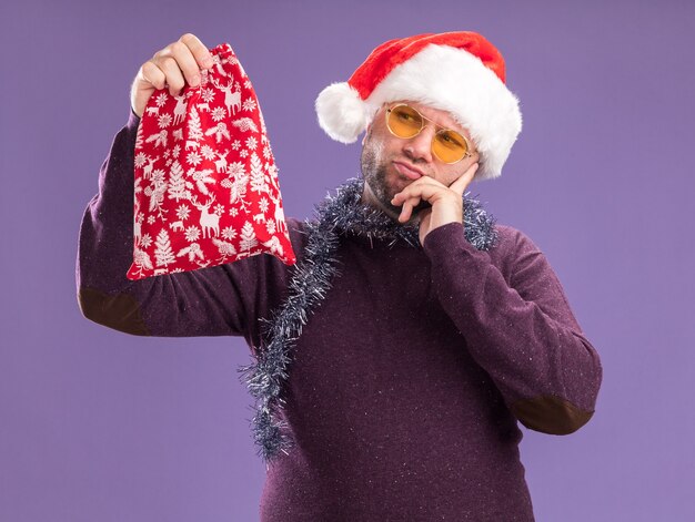 Uomo di mezza età confuso che indossa il cappello della santa e la ghirlanda di orpelli intorno al collo con gli occhiali che tengono e guardando il sacco del regalo di Natale tenendo la mano sul mento isolato sul muro viola