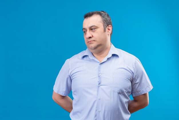 Uomo di mezza età confuso nel pensiero della camicia a strisce blu mise le mani sulla schiena su uno spazio blu