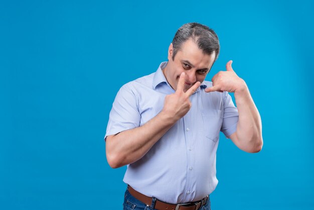 Uomo di mezza età eccitato e sorridente in camicia blu che mostra due dita e chiamami segno su uno spazio blu