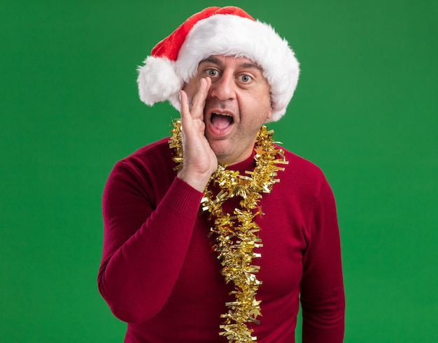 uomo di mezza età emotivo che indossa il cappello di Babbo Natale con orpelli intorno al collo gridando con la mano vicino alla bocca in piedi su sfondo verde