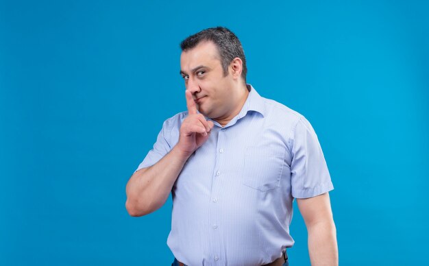 Uomo di mezza età emozionante in camicia blu che mostra il dito del segno di shhh alle labbra su uno spazio blu