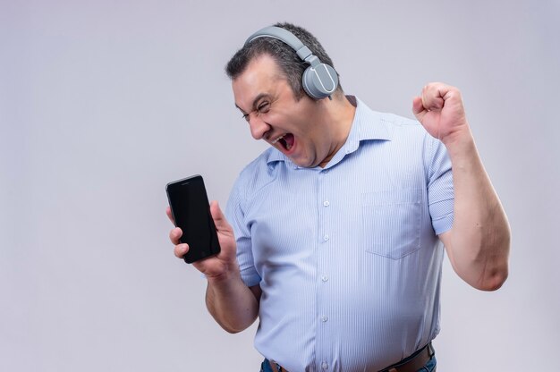 Uomo di mezza età in camicia a righe blu con la faccia urlante che indossa le cuffie che mostrano il suo smartphone su uno sfondo bianco