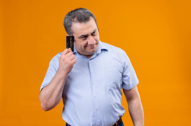 Uomo di mezza età in camicia a righe blu pensando a piani con carta di credito su uno sfondo arancione