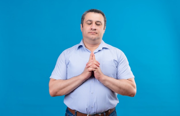 Uomo di mezza età in camicia a righe blu tenendo gli occhi chiusi in piedi e tenendosi per mano in posizione di preghiera su uno sfondo blu