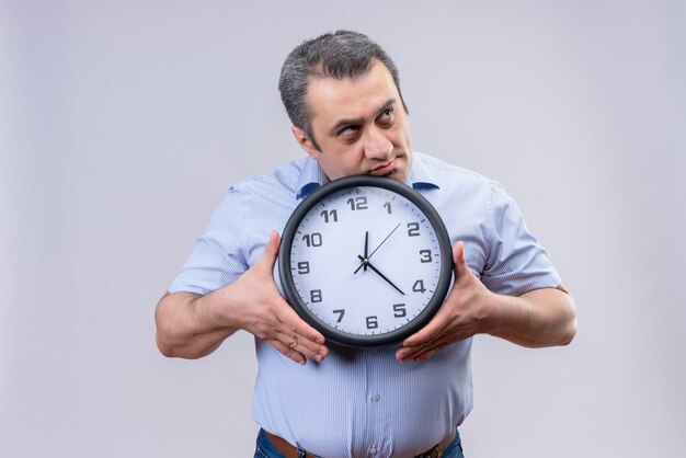 Uomo di mezza età in camicia a righe verticali blu che tiene l'orologio da parete nelle mani premuroso sull'idea confusa su una priorità bassa bianca