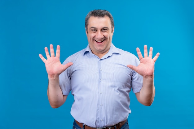 Uomo di mezza età in camicia a righe verticali blu con faccia felice che mostra e rivolto verso l'alto con il dito numero dieci su sfondo blu