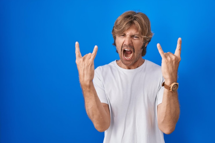 Foto gratuito uomo di mezza età in piedi su sfondo blu che grida con un'espressione pazza facendo il simbolo del rock con le mani in alto il concetto di musica pesante della star della musica