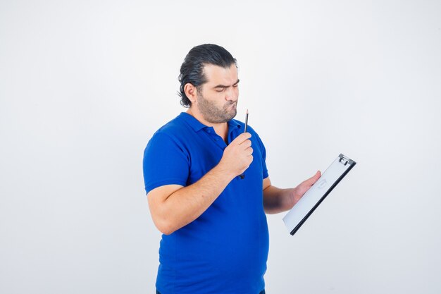 Uomo di mezza età in t-shirt polo guardando attraverso gli appunti tenendo la matita e guardando concentrato, vista frontale.