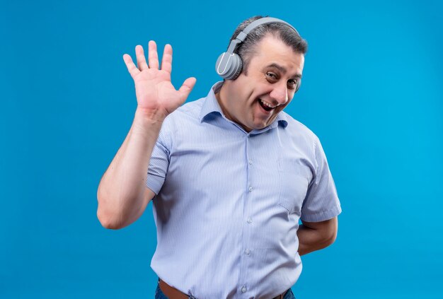 Uomo di mezza età positivo e gioioso in camicia a strisce blu che indossa le cuffie che mostrano il gesto di cinque su uno sfondo blu