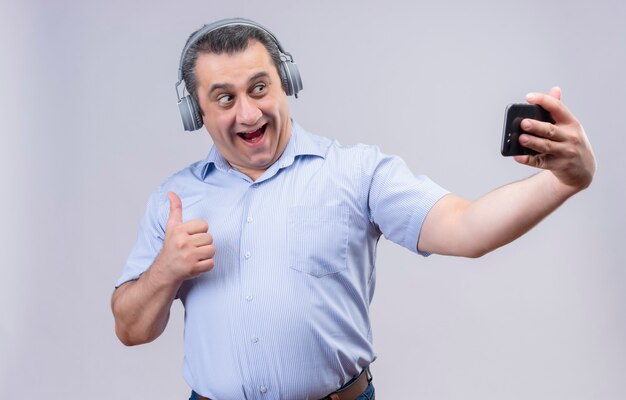 Uomo di mezza età positivo in camicia a righe verticali blu che indossa le cuffie che ride e che fa selfie sul suo telefono su una priorità bassa bianca