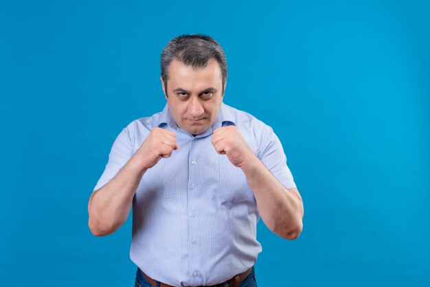 Uomo di mezza età serio e arrabbiato che indossa la camicia a strisce verticali blu che pratica i movimenti di boxe su una priorità bassa blu