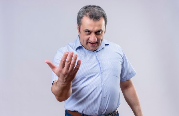Uomo di mezza età serio in camicia a strisce blu che chiama più vicino con il gesto della mano su una priorità bassa bianca