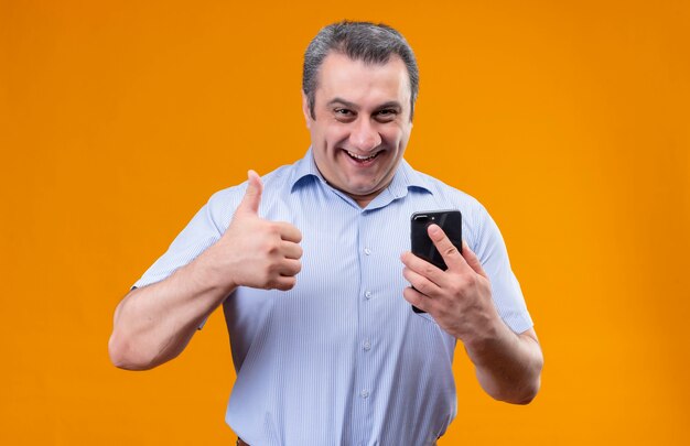 Uomo di mezza età sorridente e positivo che indossa la camicia spogliata blu che tiene il telefono cellulare e che mostra i pollici in su mentre levandosi in piedi su una priorità bassa arancione