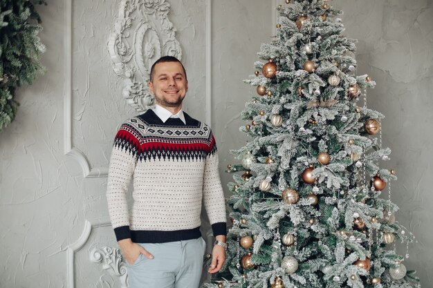 Uomo di mezza età sorridente in maglione con ornamenti in piedi accanto a un albero di Natale decorato