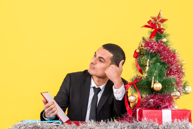 Uomo di vista frontale in vestito che si siede al tavolo guardando a destra facendo pollice in alto segno albero di Natale e regali