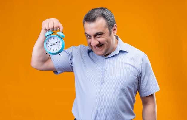 Uomo divertente e positivo in camicia a strisce blu che tiene sveglia blu e che mostra il tempo stando in piedi