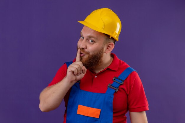 Uomo divertente giovane barbuto costruttore in uniforme da costruzione e casco di sicurezza che fa gesto di silenzio con il dito sulle labbra su sfondo viola