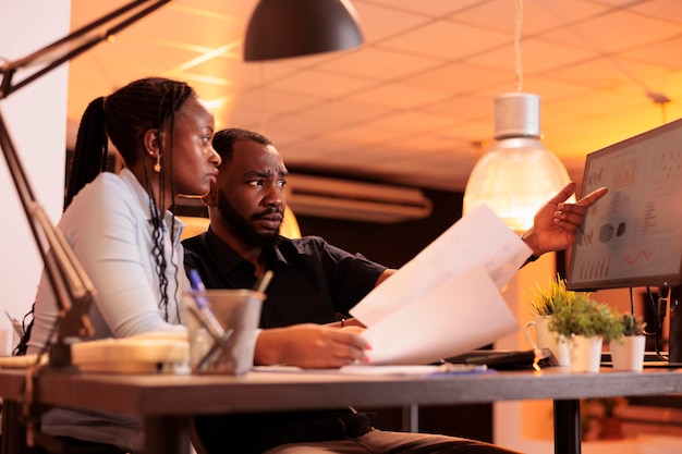 Uomo e donna afroamericani che collaborano al lavoro di squadra per fare un rapporto di presentazione, guardando i dati su computer e documenti. Lavorare insieme alle scartoffie in ufficio durante il tramonto.