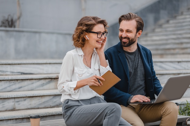 Uomo e donna attraenti seduti sulle scale nel centro urbano, lavorando insieme sul laptop