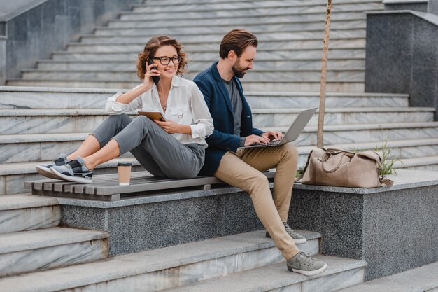 Uomo e donna attraenti seduti sulle scale nel centro urbano, lavorando insieme sul laptop