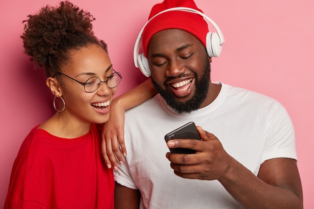 Uomo e donna etnici felici guardano video divertenti su smartphone, uomo nero con cappello rosso e maglietta bianca, indossa le cuffie, mostra la nuova app alla ragazza