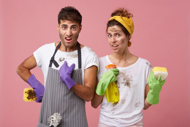 Uomo e donna insoddisfatti in vestiti sporchi che tengono la spugna e lo spruzzo di lavaggio che sono disordinati