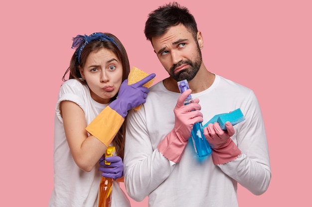 Uomo e donna scontenti infelici tengono spray, spugne, vestiti con abiti casual bianchi, coinvolti nella pulizia, fanno le faccende domestiche nei fine settimana