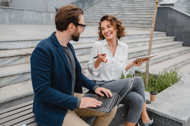 Uomo e donna sorridenti attraenti che parlano seduti sulle scale nel centro urbano, prendendo appunti