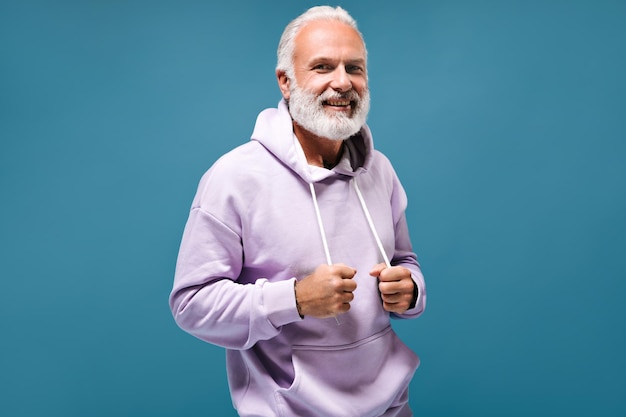 Uomo elegante con felpa con cappuccio bianca sorridente Ragazzo adulto alla moda dai capelli grigi con barba con tatuaggi in felpa lilas che guarda nella fotocamera