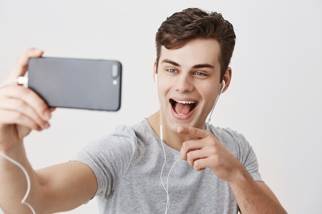 Uomo europeo bello che tiene telefono cellulare, che posa per il selfie, che fa videochiamata, ampiamente sorridente, indicando con il dito indice allo schermo del telefono cellulare. Comunicazione e tecnologia moderne.