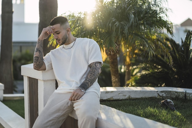 Uomo europeo sconvolto con una camicia bianca con le braccia tatuate seduto all'aperto
