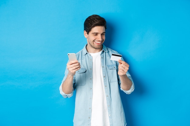 Uomo felice bello che paga per qualcosa in linea, tenendo la carta di credito e il telefono cellulare, acquisto in internet, in piedi su sfondo blu.