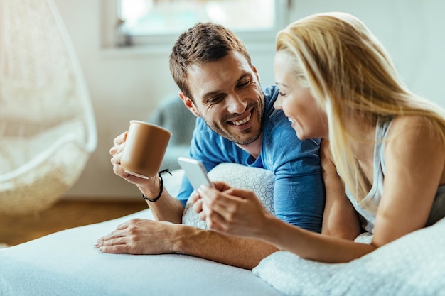 Uomo felice che beve il caffè del mattino e comunica con la sua ragazza che sta usando il telefono cellulare in camera da letto