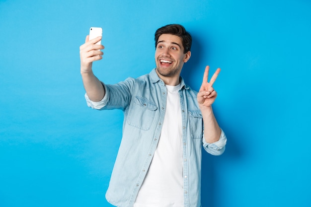 Uomo felice che prende selfie e mostra il segno di pace su sfondo blu, tenendo il telefono cellulare
