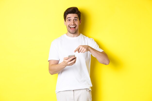 Uomo felice che punta allo smartphone per mostrare promo, controlla l'offerta internet, in piedi sopra il giallo