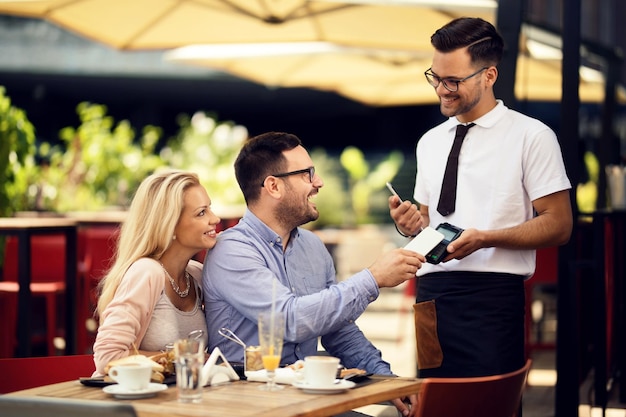 Uomo felice che usa lo smartphone e paga un conto a un cameriere mentre è con la sua ragazza in un ristorante