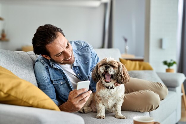 Uomo felice che usa lo smartphone mentre ci si rilassa sul divano con il suo cane che sta sbadigliando