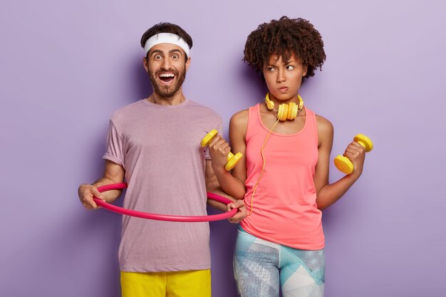 Uomo felice in abbigliamento casual, altalene hula hoop, donna dalla pelle scura alza le braccia con manubri, vestito con abbigliamento sportivo, fa diversi esercizi fisici, isolato sopra il muro viola. Sport e allenamento