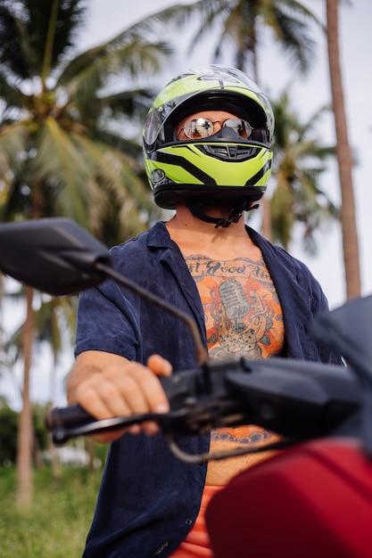 Uomo forte tatuato sul campo della giungla tropicale con una moto rossa