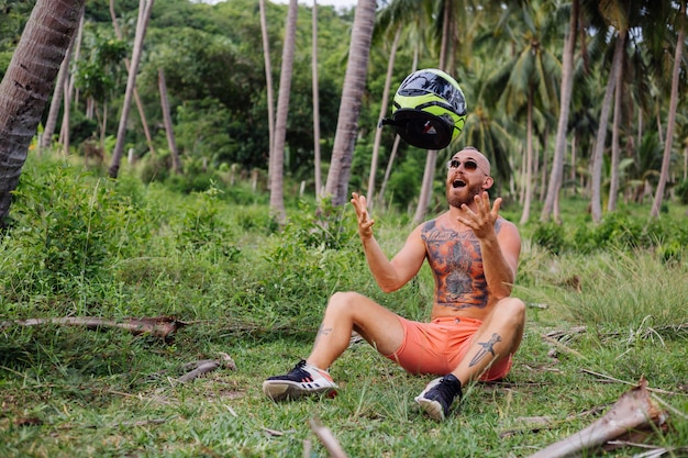 Uomo forte tatuato sul campo della giungla tropicale con una moto rossa