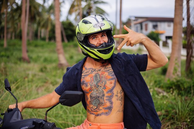 Uomo forte tatuato sul campo della giungla tropicale con una moto rossa