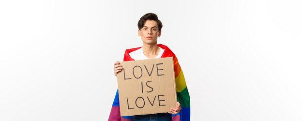 Uomo gay serio e fiducioso che indossa la bandiera arcobaleno lgbt con cartello per la parata dell'orgoglio in piedi