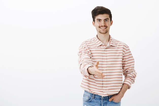 Uomo gentile e pulito che saluta il nuovo datore di lavoro. Ritratto di bello modello maschio amichevole fiducioso con baffi e barba, tirando la mano verso la stretta di mano, accogliendo il nuovo arrivato oltre il muro grigio